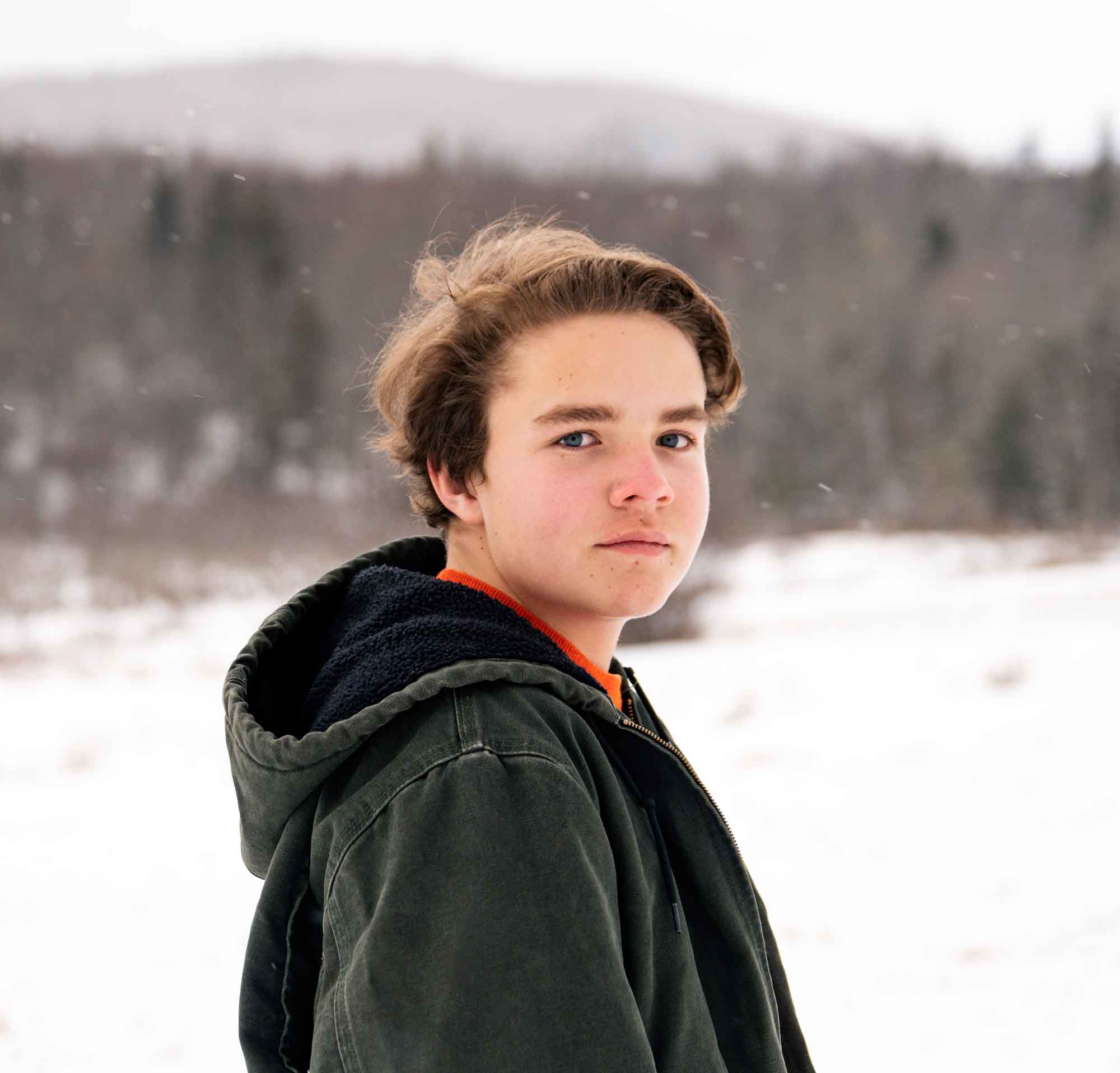 A young man standing in a field wearing a coat while it gently snows.